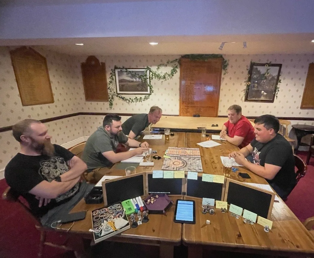 Adults gathered around a tabletop roleplaying game in a pub, with the shared map and Host setup visible